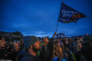 Crowd of festival-goers at dusk, a person flags a dark blue flag that reads 'Common Kings' above the crowd.
