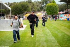 Two men run across a grassy field at an outdoor festival, tents and spectators in the background.