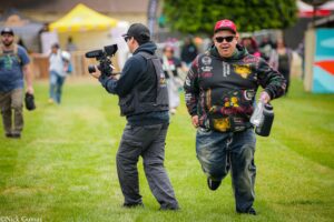 Videographer with a shoulder rig and microphone filming a man in a colorful hoodie at an outdoor event.