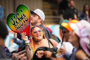 Man kissing a large rainbow heart sign at a crowded parade, with colorful accessories in the foreground.