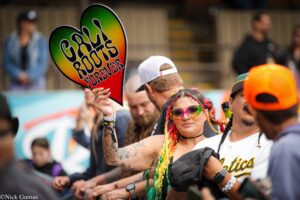 A person holds a rainbow-colored sign that reads 'Good Vibes' at a crowded outdoor event or festival. In the foreground, a woman with colorful braids and pink sunglasses smiles amid the crowd, with others in the background.