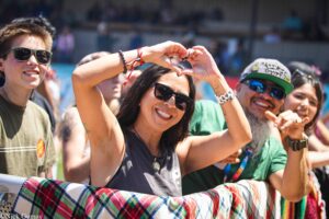 Group of smiling people at an outdoor event, forming a heart shape with their arms, sunny day, wearing sunglasses and casual clothes.