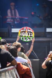 Audience member raises a colorful circular sign that reads 'CALI ROOTS FEST' at a concert, with a blurred musician on stage in background.