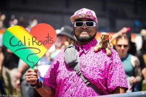 Parade participant wearing a pink leopard-print shirt and white sunglasses, holding a rainbow heart sign and a small plush toy.