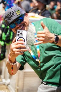 Smiling man wearing a gray cap with 'Pacific Roots' and blue mirrored sunglasses, taking a selfie with a smartphone at an outdoor event.