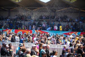 Crowd at an outdoor festival behind a railing with colorful mural on the barrier and confetti falling from above.