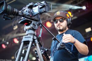 Cameraman wearing sunglasses and a cap operates a large professional video camera on a tripod at a stage event with headphones on.
