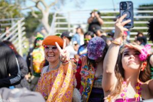 Smiling woman in an orange hat and white sunglasses flashes a peace sign at a colorful outdoor festival crowd.