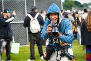 Photographer in blue hoodie smiling while holding a Nikon camera with a long lens, crowd in the background at an event.