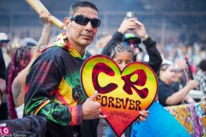 Man wearing sunglasses at a crowded outdoor event, holding a large rainbow-colored heart sign in the foreground.