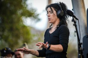 Outdoor performance: a woman with dark hair wearing large over-ear headphones, gesturing with her hands on a stage next to equipment.