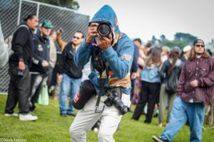 Photographer in a blue hoodie aiming a large camera at an outdoor crowd during an event.