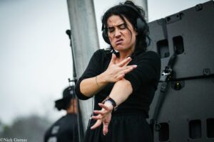 Female performer on stage wearing headphones, making an expressive hand gesture with a focused, intense expression.