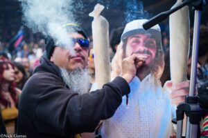 Man with a beard and sunglasses leans close to a large poster of a smiling woman at a crowded outdoor event, smoke curling from a cigarette nearby.