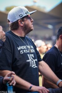 Side profile of a man in a black T-shirt and white mesh cap at an outdoor event, wearing blue earphones and a wristband.