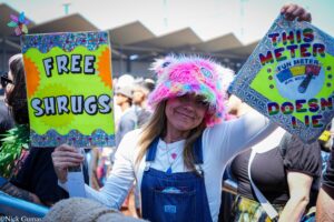 Woman in a rainbow fuzzy hat smiles while holding a bright sign that reads 'FREE SHRUGS' at a crowded outdoor event in sunlight.
