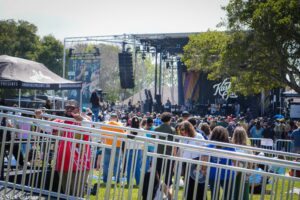 Outdoor concert scene with a large stage, speakers, and a crowd behind metal barriers on a sunny day.