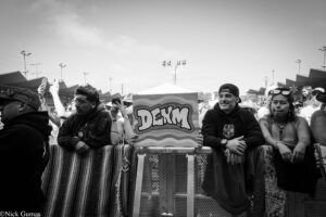 Crowd at an outdoor event behind a barrier; a sign with graffiti lettering 'DEKM' is center, with people taking photos nearby (black and white).