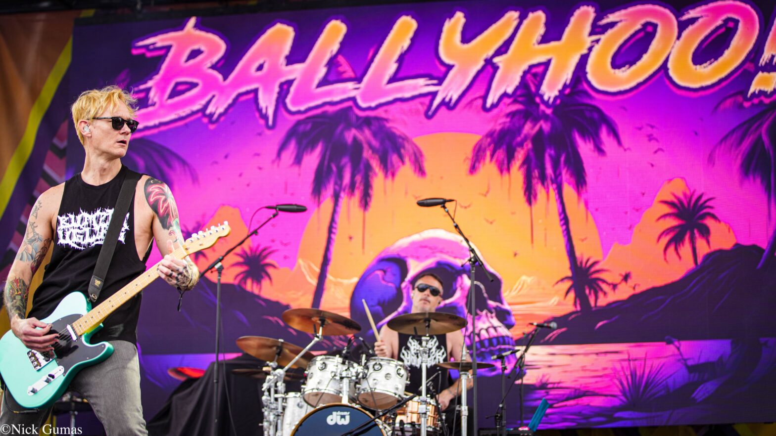 Guitarist with blond hair and sunglasses plays a mint-colored electric guitar on a vibrant stage backdrop with palm trees and a sunset scene behind the drummer.