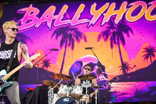Guitarist with blond hair and sunglasses plays a mint-colored electric guitar on a vibrant stage backdrop with palm trees and a sunset scene behind the drummer.