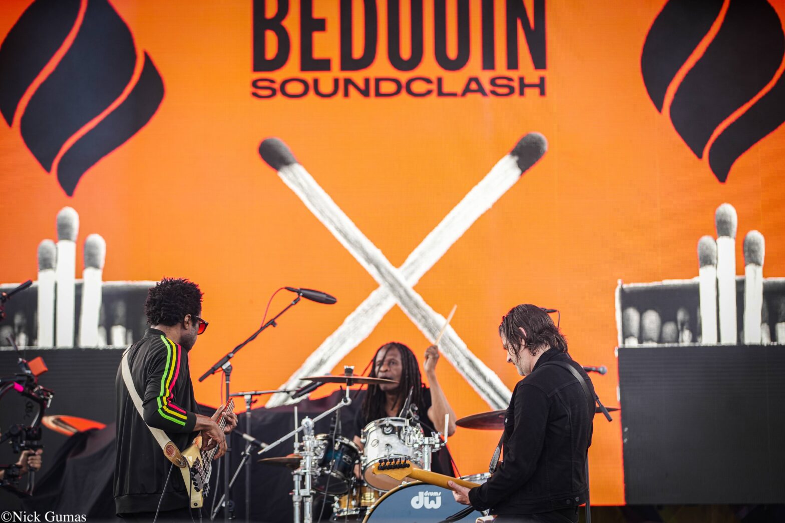 Live band performing on an outdoor stage with a bright orange backdrop reading 'BEDOOIN SOUNDCLASH'; guitarist left, drummer center, guitarist right, microphones and drums visible.