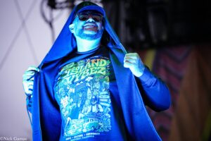 Male performer on stage in blue lighting, unzipping hoodie to reveal a colorful illustrated concert T‑shirt and wearing sunglasses.