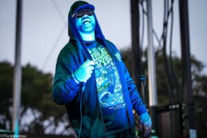 Male performer on a stage under blue lighting, wearing a hoodie and sunglasses, singing into a microphone.