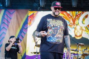 Male performer wearing a black graphic T-shirt, cap and sunglasses on a colorful mural stage, holding a microphone.