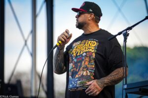Male performer singing into a handheld microphone on an outdoor stage, wearing a black cap and a colorful graphic T-shirt showing text and imagery, with tattooed arms visible.