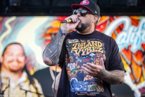 Male performer singing into a microphone on stage, wearing sunglasses and a cap with tattoos visible on his arm, colorful shirt backdrop behind him.