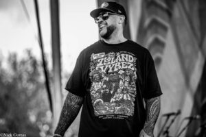 Black-and-white portrait of a tattooed man wearing a cap and sunglasses, smiling outdoors in a graphic festival T-shirt.
