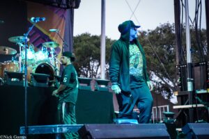Male performer in a green hoodie and sunglasses sings on an outdoor stage, with a drummer and keyboardist visible in the background.