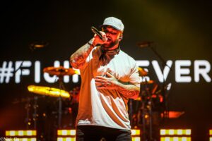 Male singer wearing a gray cap and white t-shirt sings into a microphone, tattoos visible on his arms, on a dimly lit stage.