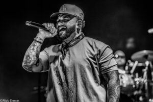 Black-and-white photo of a male singer performing on stage, gripping a handheld microphone, wearing a cap and bandana.