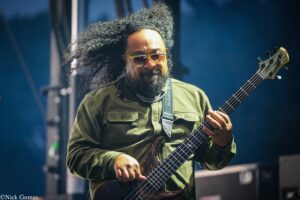 Bass guitarist performing on stage, wearing a green shirt and yellow sunglasses, with long curly hair and a dark bass guitar.
