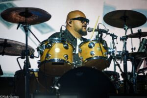 Drummer wearing sunglasses performs behind a brown drum kit with cymbals on stage, mic in front of him