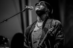 Black-and-white photo of a male singer wearing sunglasses and a cap, singing into a microphone on stage.