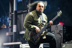 Bassist performing live on stage, wearing a green jacket and sunglasses, focused on playing a dark wood bass guitar.