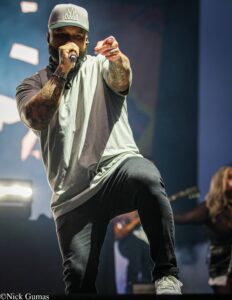 Male singer on stage, wearing a gray cap with a crown logo, tattoos showing, holding a microphone and pointing toward the audience.