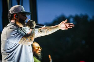 Male vocalist on stage wearing a gray cap and patterned bandana, singing into a handheld microphone with his tattooed forearm extended toward the audience.