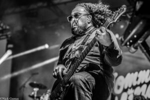 Black-and-white photo of a bass guitarist on stage, wearing glasses and a beard, deeply focused while playing.