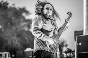 Bassist with long curly hair and glasses playing an electric bass on a stage during a live performance.