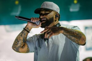 Male singer wearing a gray cap and gray shirt, singing into a handheld microphone on stage, with visible tattoos on both arms.