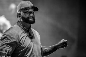 Male performer on stage, wearing a cap, bandana, and visible tattoos on the arm, captured in black and white.
