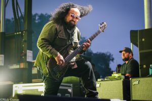 Bass guitarist with long curly hair and sunglasses plays on an outdoor stage in a green jacket.