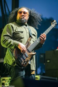 Bass guitarist performing on stage, wearing green shirt and sunglasses, playing a wooden five‑string bass with a strap and curly hair.