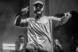 Male performer singing into a microphone on stage, wearing a cap and bandana, with tattoos on his arms (black-and-white).