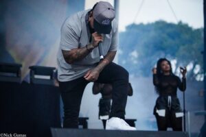 Male rapper on stage, microphone in hand, wearing a gray shirt and cap, performing with tattoos visible on arms, smoke effects in background.