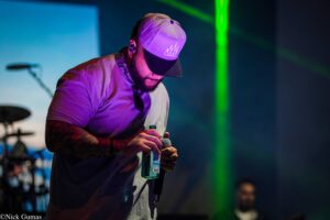 Male singer on stage under purple lighting, wearing a light shirt and purple cap with a crown logo, holding a microphone and a bottle of drink.