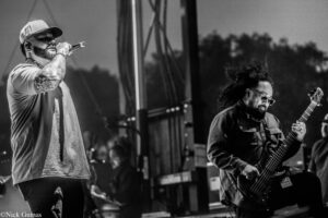 Male vocalist with a cap and tattooed arm sings into a microphone on stage, while a guitarist with sunglasses plays nearby on a black-and-white concert photo.
